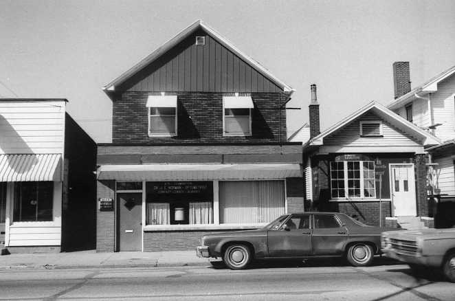 A black and white photo of a vintage car parked by a building with a steep gable roof.