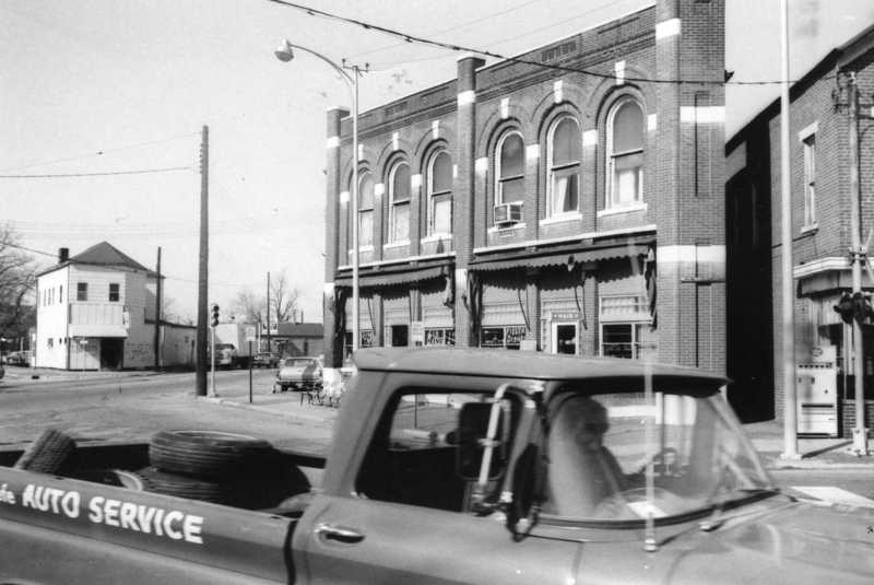 Black and white photo of an old street scene with a classic car in the foreground and vintage buildings in the background.