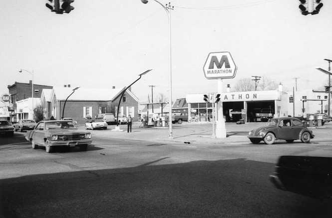 Black and white photo of an intersection with vintage cars, Marathon gas station, and street signs.
