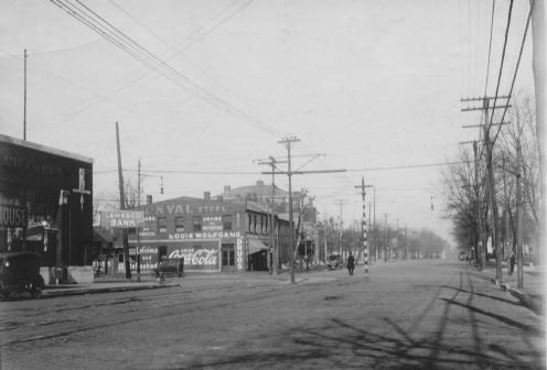 A vintage street scene with buildings, advertisements like Coca-Cola, utility poles, and a few pedestrians. Black and white photo.