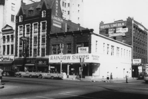 Black and white photo of a city street with old buildings, signs for "Hotel Lincoln," "Rainbow Shops," and pedestrians walking.