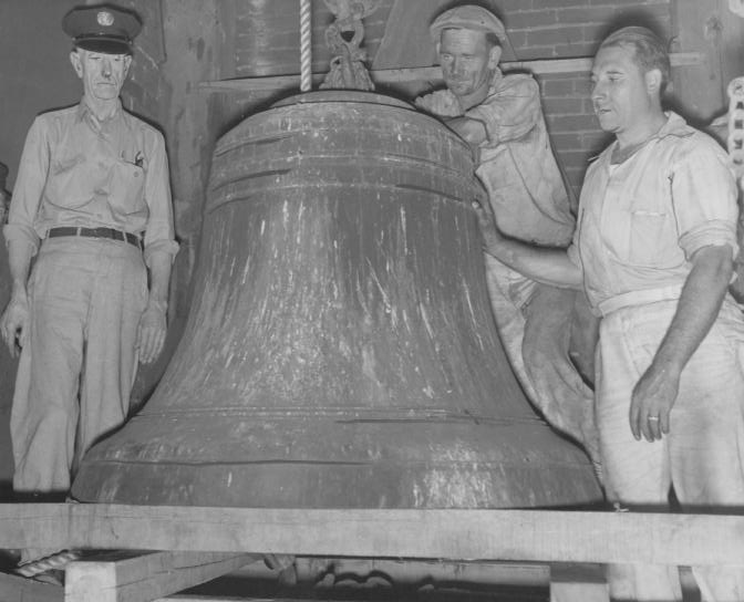 A large bell with two men, one in a cap and one in a sleeveless shirt, possibly in a bell tower. Black and white photo.