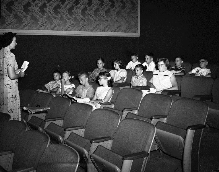 A woman stands speaking to a group of seated children in a vintage classroom setting with chairs and patterned wallpaper.