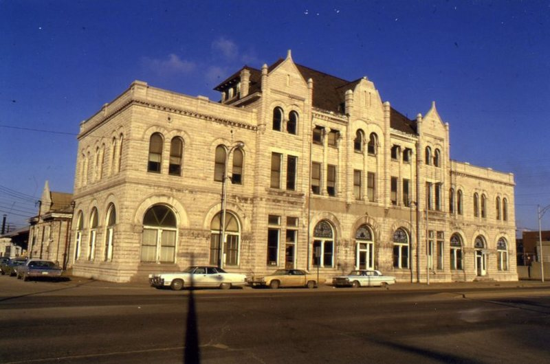 An ornate, historic stone building with arched windows on a sunny day, cars parked alongside the street.