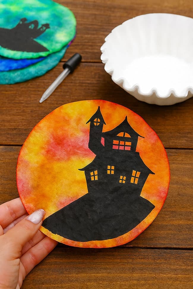 A hand holds a circular, colorful art piece with a silhouetted spooky house against a vibrant background.