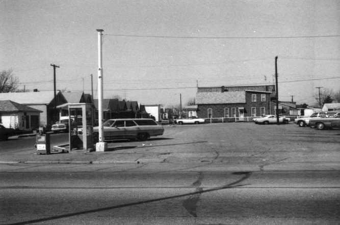 A vintage black and white photo of a street scene with cars, buildings, and a gas station.
