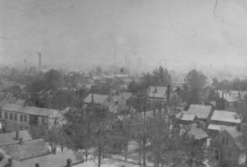 Old black and white photo of a residential area with houses and bare trees, possibly winter, with an industrial backdrop.