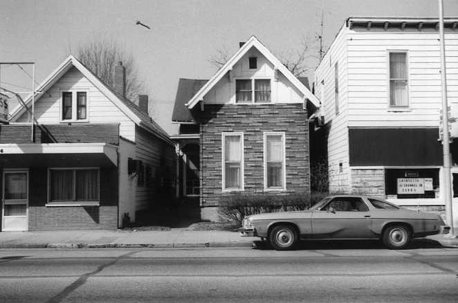 Black and white photo of two houses with a classic car parked in front and a bird flying overhead.