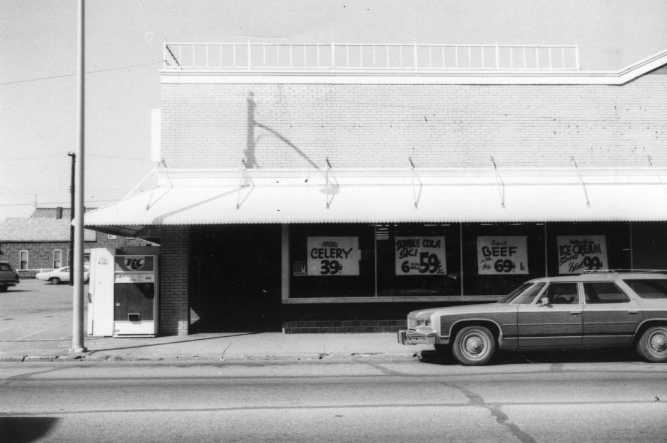 Black and white photo of a vintage storefront with produce ads, a payphone, and an old car parked out front.