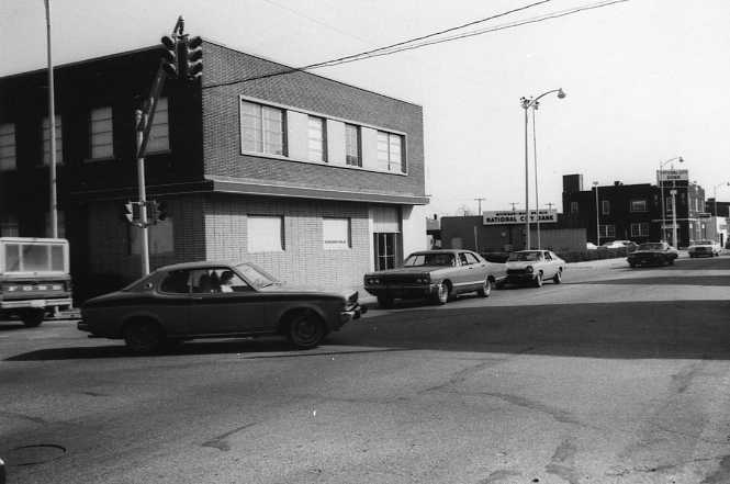 Black and white photo of a street corner with vintage cars and buildings.