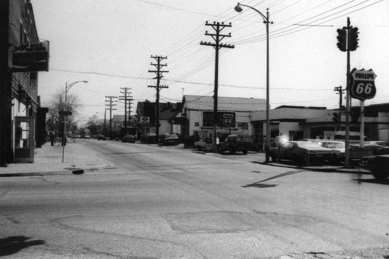 Black and white photo of a vintage street with cars, Route 66 sign, utility poles, and buildings.