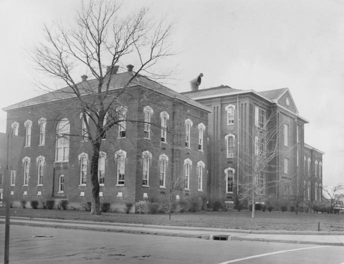 An old, two-story brick building with white-trimmed windows, bare tree branches in the foreground, against a clear sky.