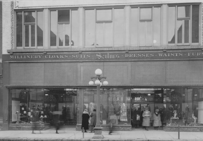 Vintage photo of a storefront advertising millinery, cloaks, suits, with people passing by and examining display windows.