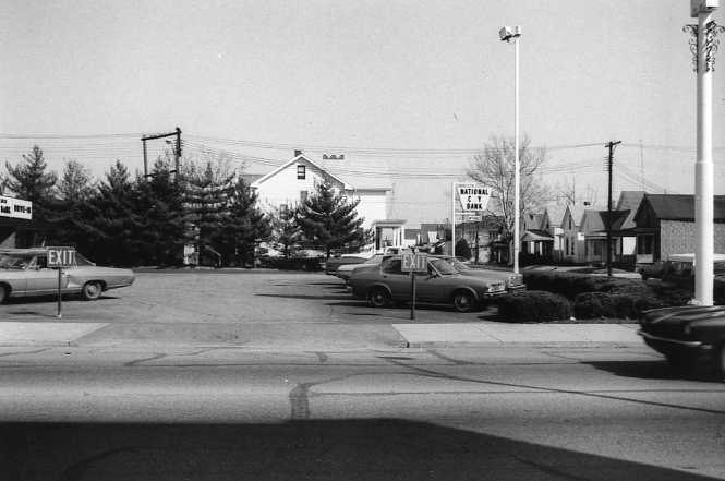 Black and white photo of a parking lot with vintage cars, buildings in the background, and exit signs.