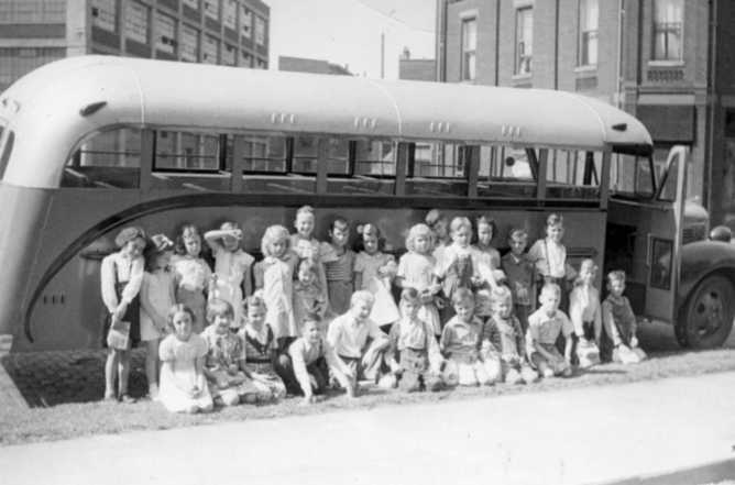 A group of children posing in front of an old-style bus on a sunny day, with buildings in the background.