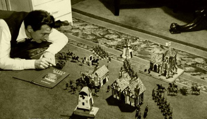 Actor Peter Cushing is laying on the floor, playing with model soldiers and buildings, possibly engaged in a tabletop game or diorama setup.