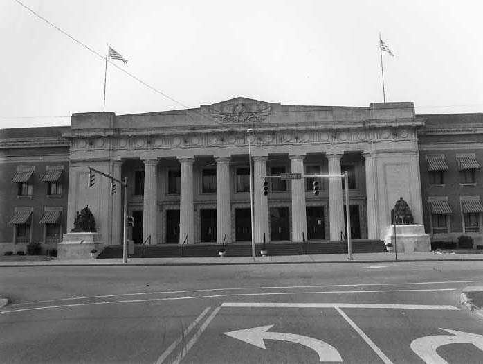 A black and white photo of a classical building with columns, statues, and flags, viewed from across a street with directional arrows.