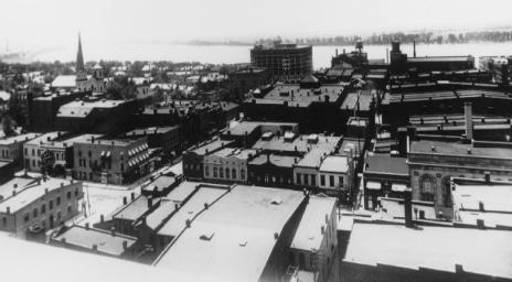 Historic black and white photo of an old cityscape with various buildings and a view of a water body in the distance.