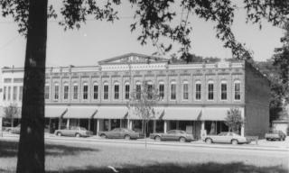 A black and white photo of a historic two-story building with parked cars. Trees partially obscure the view.