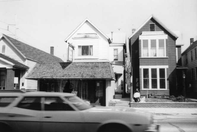 Black and white photo of a residential street with houses and a moving car in the foreground.