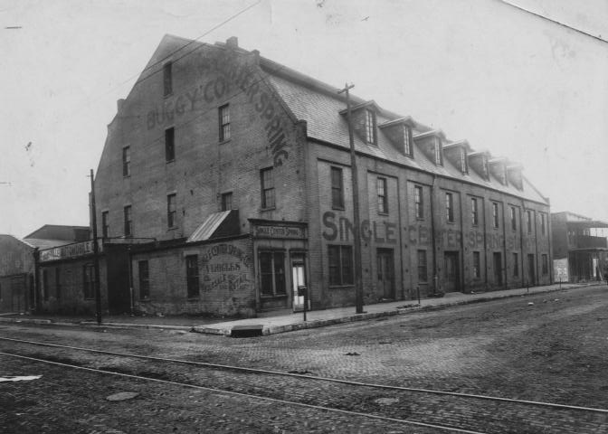 Vintage photo of an old brick building with "Buggy Co Springs" sign, near cobblestone streets with tram tracks.