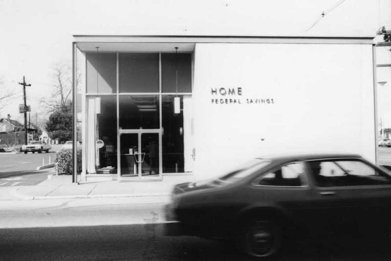 A black and white photo of a building with "HOME FEDERAL SAVINGS" sign and a blurred car passing by.