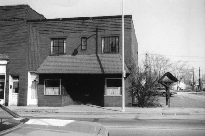 Black and white photo of a two-story building with a car in the foreground and a stop sign on the right.