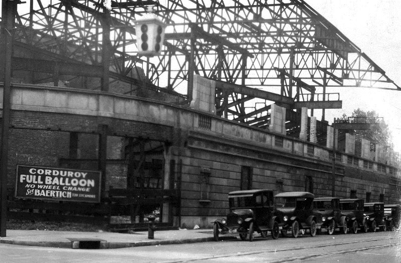 Old photo of a street with vintage cars, an advertisement on a wall, and a steel structure overhead, likely a bridge or train tracks.