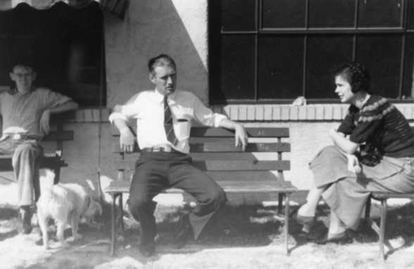 Three people and a dog outside near a bench, one person is standing. Vintage black and white photo.