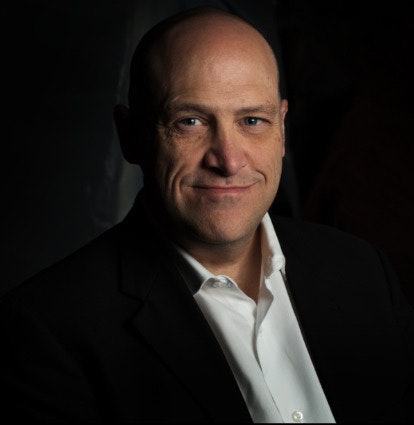Portrait of a smiling bald man in a suit with a white shirt, dark background.