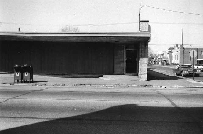 Black and white photo of a street with buildings, cars, and shadows.