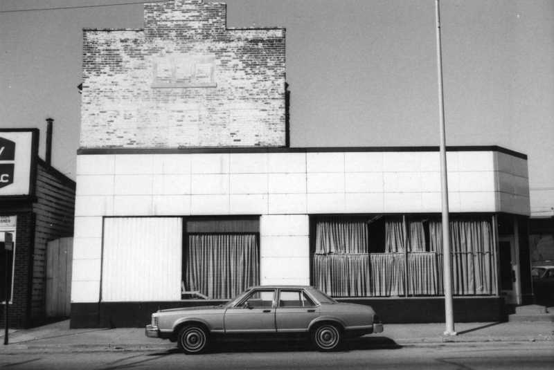 A vintage car parked in front of an old building with boarded-up windows, under a clear sky. Black and white photo.