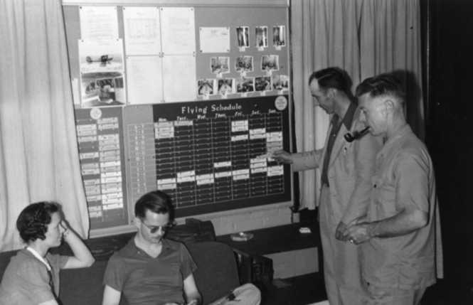 People examining a flying schedule board, with photos and notices on the wall behind them.