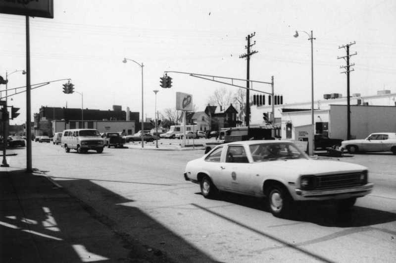Black and white photo of a busy intersection with vintage cars, traffic lights, and some commercial buildings.
