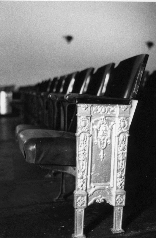 A row of ornate, empty theater seats in black and white.