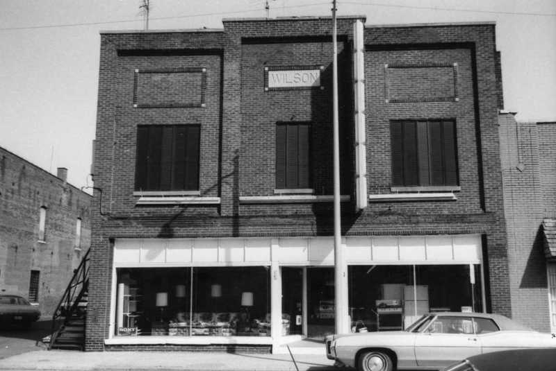 A two-story brick building with a sign "WILSON" and a vintage car parked in front.