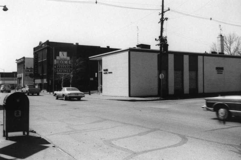 Black and white photo of a street corner, buildings, vintage cars, and a mailbox.