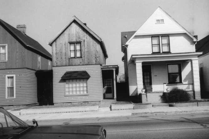 Black and white photo of two adjacent houses, one with a weathered facade, the other painted and well-maintained.
