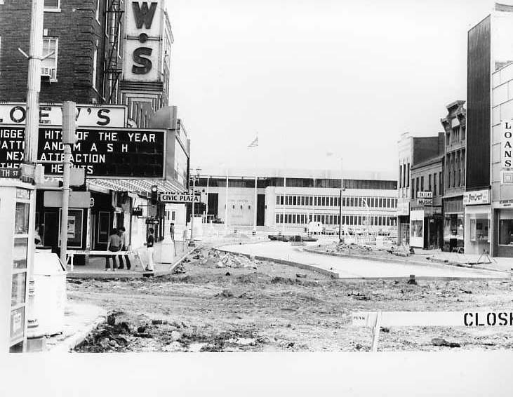 Urban construction site, torn up street, old buildings, marquee with movie titles, "Civic Plaza" in background, American flag.