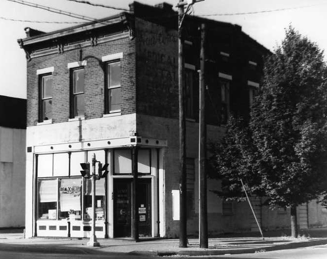 An old two-story building with a shop on the ground floor, street signs, and a tree. Black and white photo.