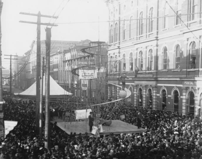 Historic black and white photo depicting a crowded event with an aircraft model, banners, and period architecture.