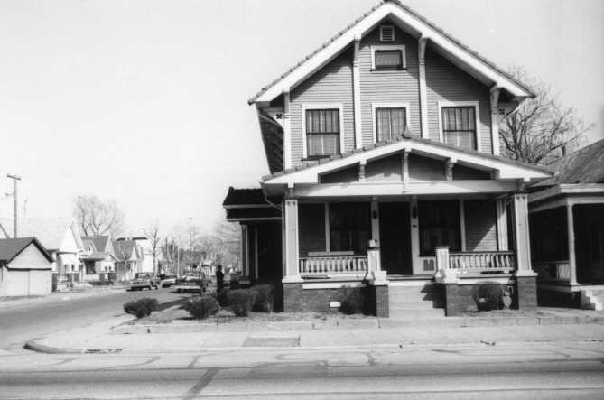 Black and white photo of a two-story house with a porch, in a residential area.