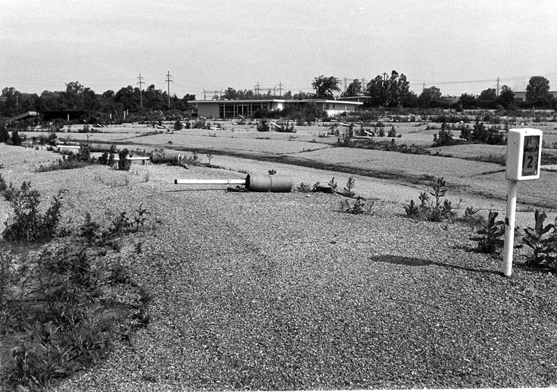 Gravel-covered area with a sign, overgrown plots, and a building in the distance.