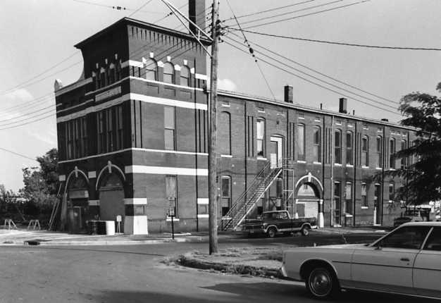 Black and white photo with vintage cars and brick buildings with arches.