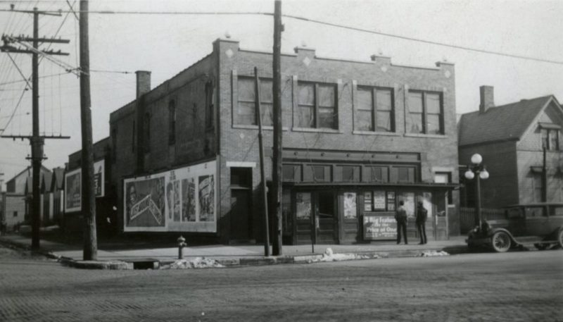 Black & white photo of an old street corner with buildings, ads, a vintage car, and a couple of people.