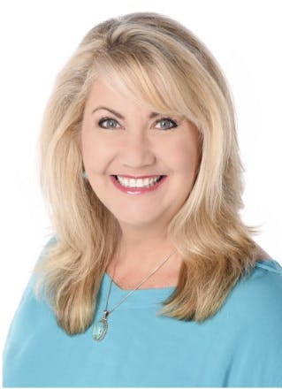 A smiling woman with blonde hair wearing a blue top and a necklace, with a white background.