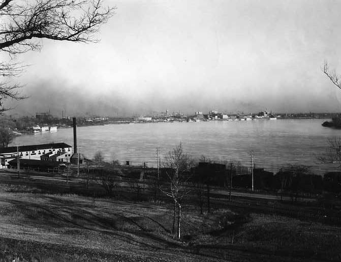 Black and white photo of a river landscape with mist, a boat dock and some industrial buildings in the distance.