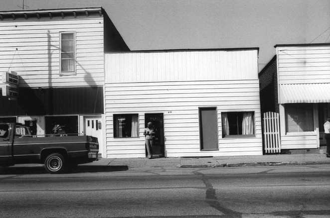 Black and white photo of a street with a truck parked outside older buildings, one with a person standing in the doorway.