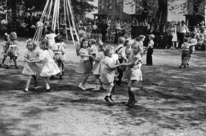 Children playing Ring Around the Rosie, with a swing set in the background, watched by spectators. Black and white, vintage feel.