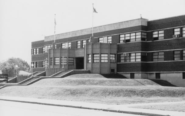 Black and white photo of a two-story brick building with flags, stairs, and landscaping.